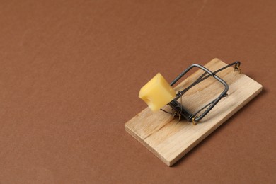 Photo of Spring-loaded bar mousetrap with piece of cheese on brown background, space for text