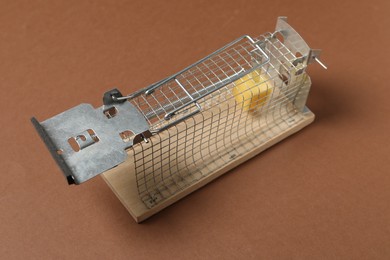 Photo of Metal mouse trap with piece of cheese on brown background, closeup