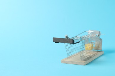 Photo of Metal mouse trap with piece of cheese on light blue background, space for text