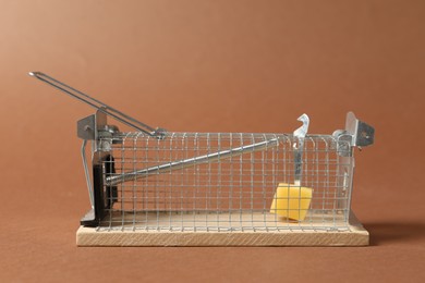 Photo of Metal mouse trap with piece of cheese on brown background