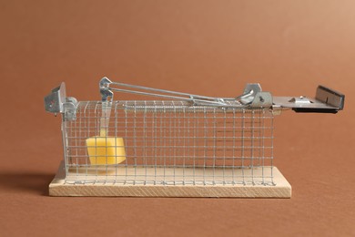 Photo of Metal mouse trap with piece of cheese on brown background