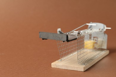 Photo of Metal mouse trap with piece of cheese on brown background, closeup. Space for text