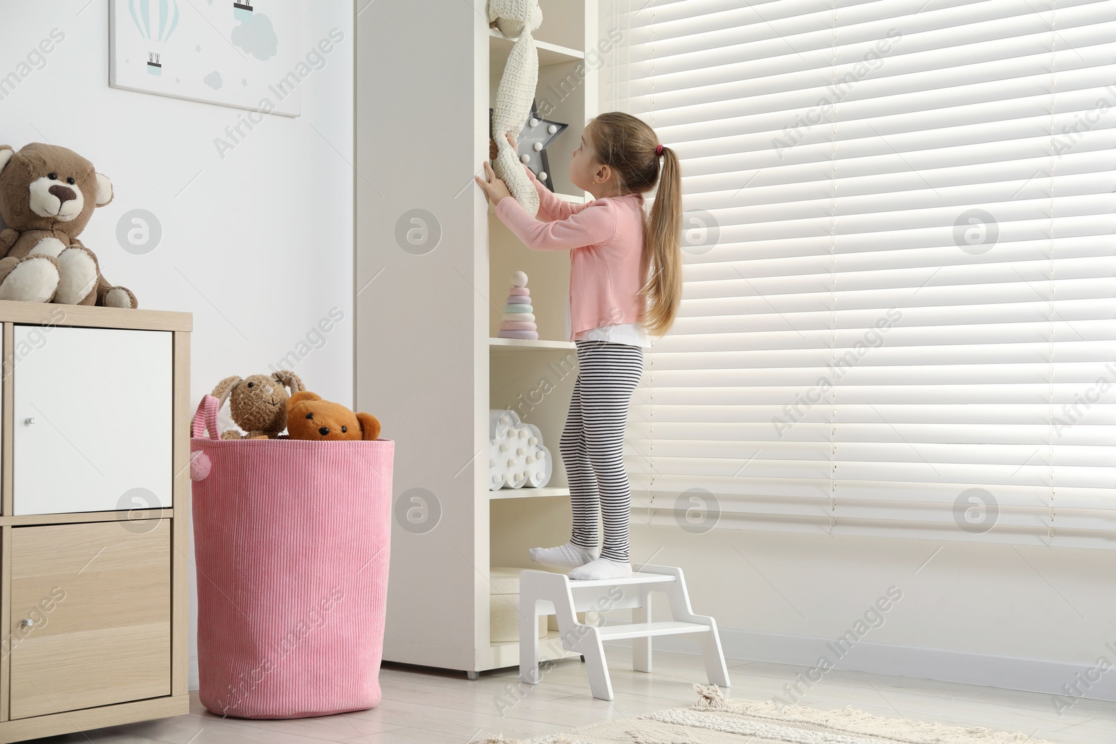 Photo of Little girl standing on step stool and reaching for toys on shelf indoors. Space for text