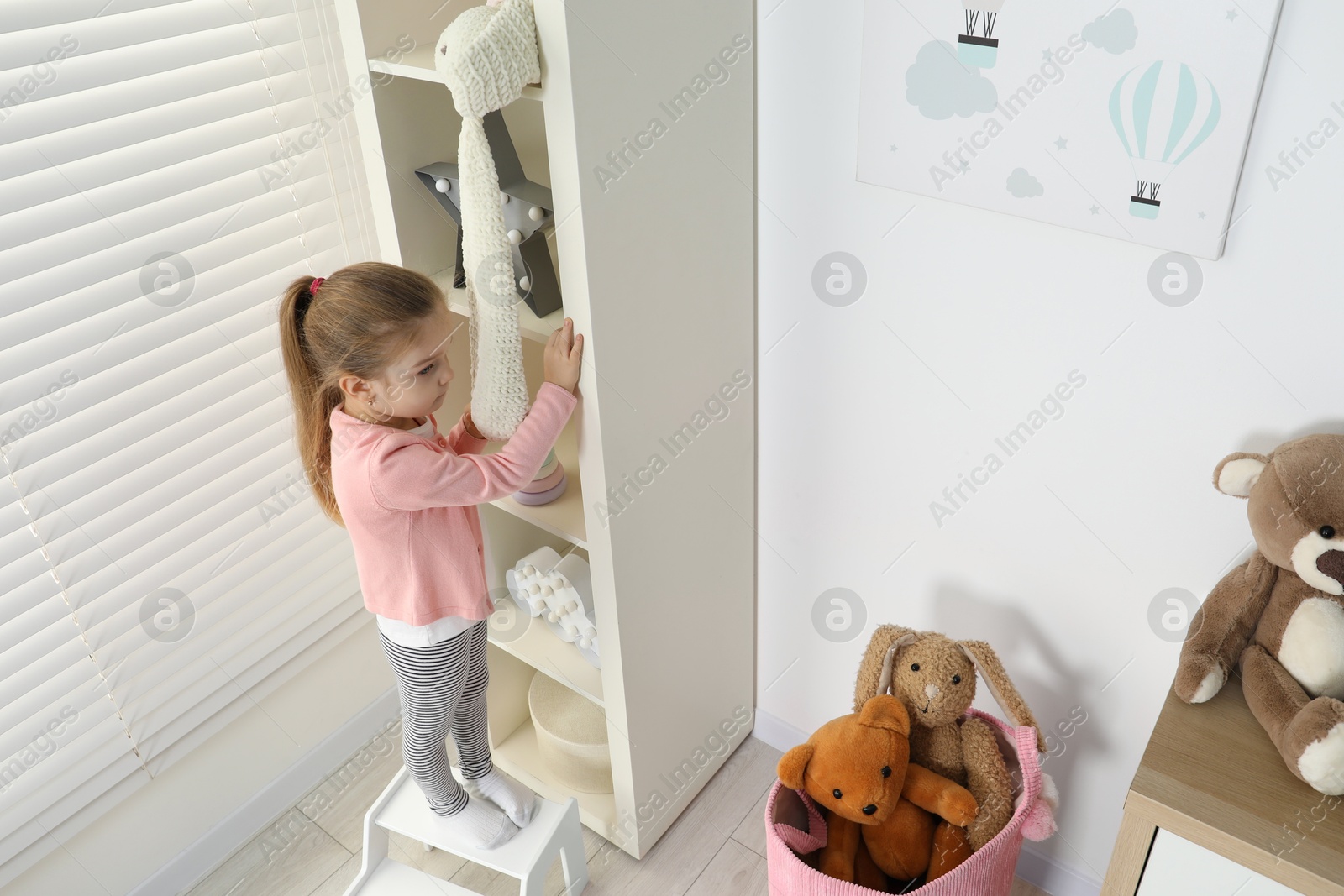 Photo of Little girl standing on step stool and reaching for toys on shelf indoors, above view