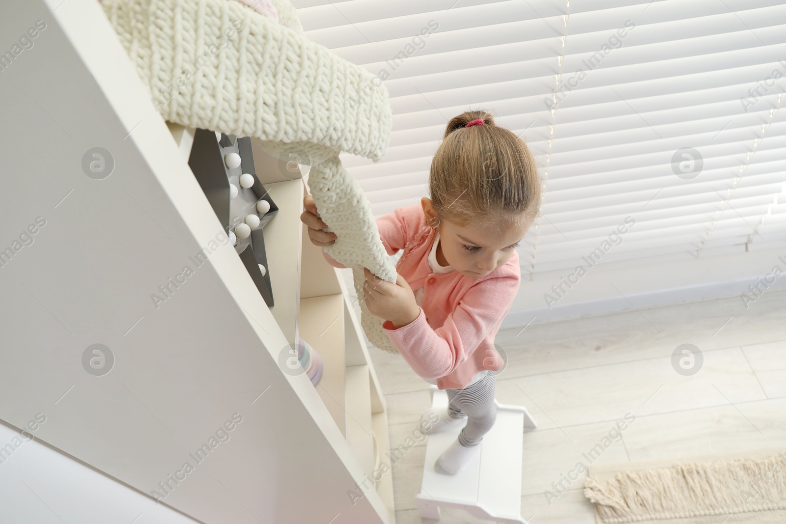 Photo of Little girl standing on step stool and reaching for toys on shelf indoors, above view