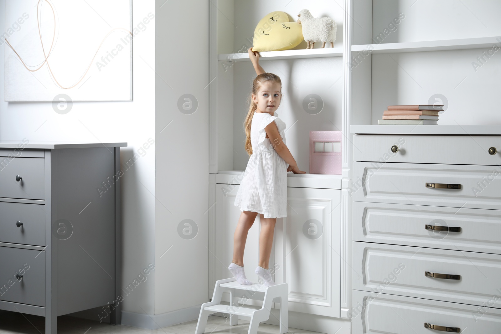 Photo of Little girl standing on step stool and reaching for toys on shelf indoors