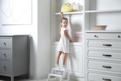 Little girl standing on step stool and reaching for toys on shelf indoors