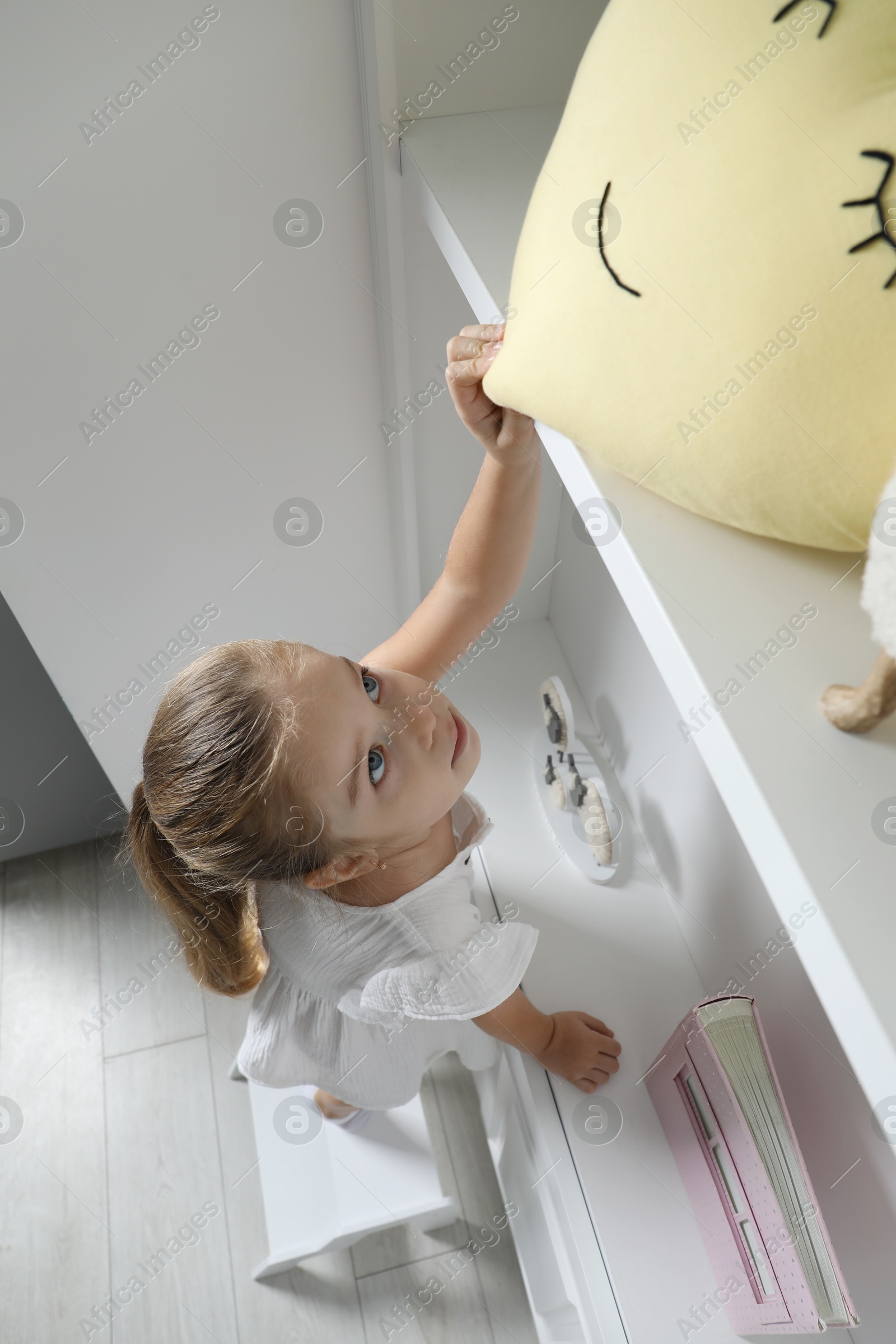 Photo of Little girl standing on step stool and reaching for toys on shelf indoors, above view