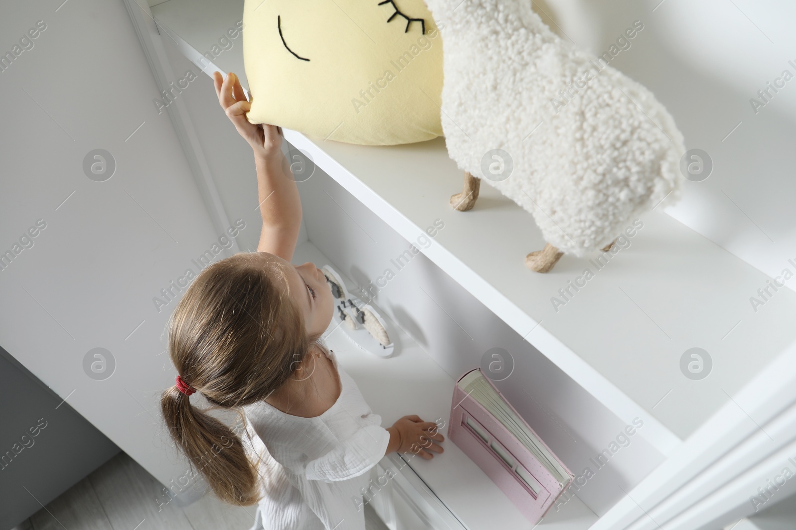 Photo of Little girl reaching for toys on shelf indoors, above view