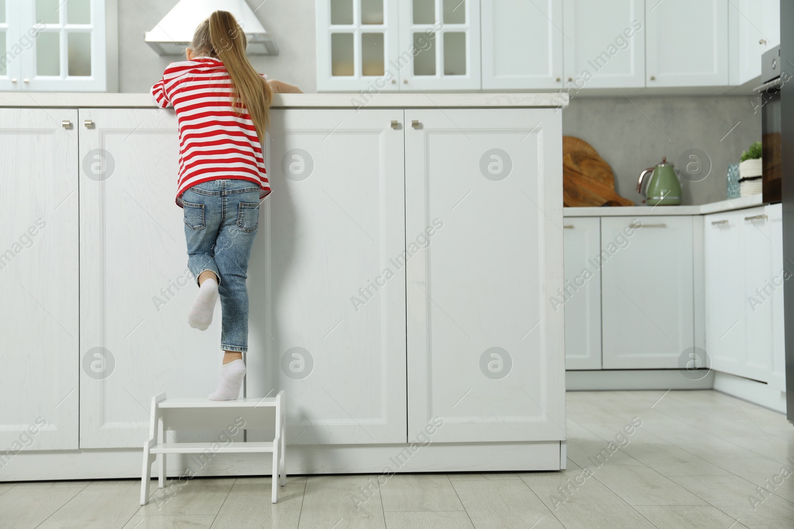 Photo of Little girl standing on step stool and reaching towards counter in kitchen, back view. Space for text