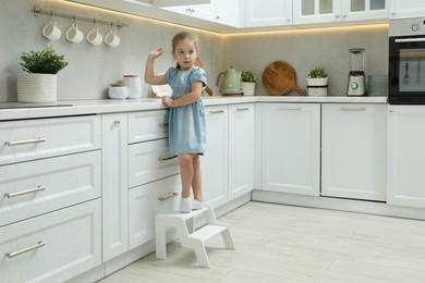 Little girl standing on step stool and reaching towards counter in kitchen