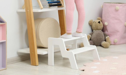 Photo of Little girl standing on step stool and reaching for toys on shelf indoors. Space for text