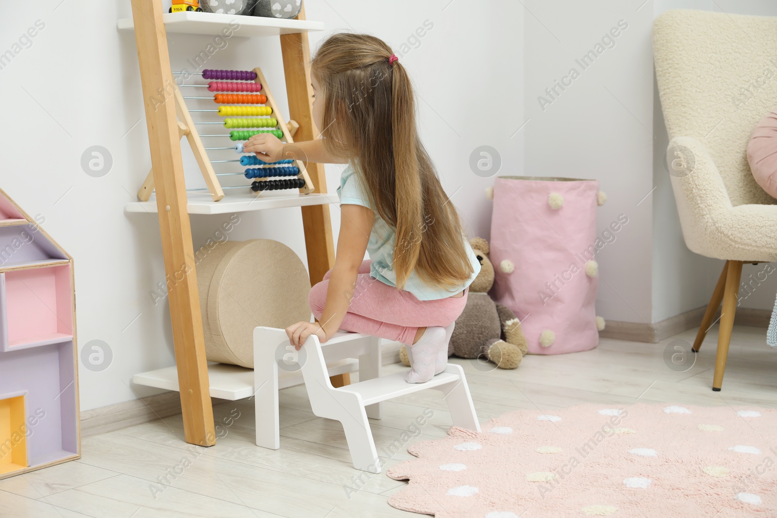 Photo of Little girl sitting on step stool near shelf with toys at home, back view