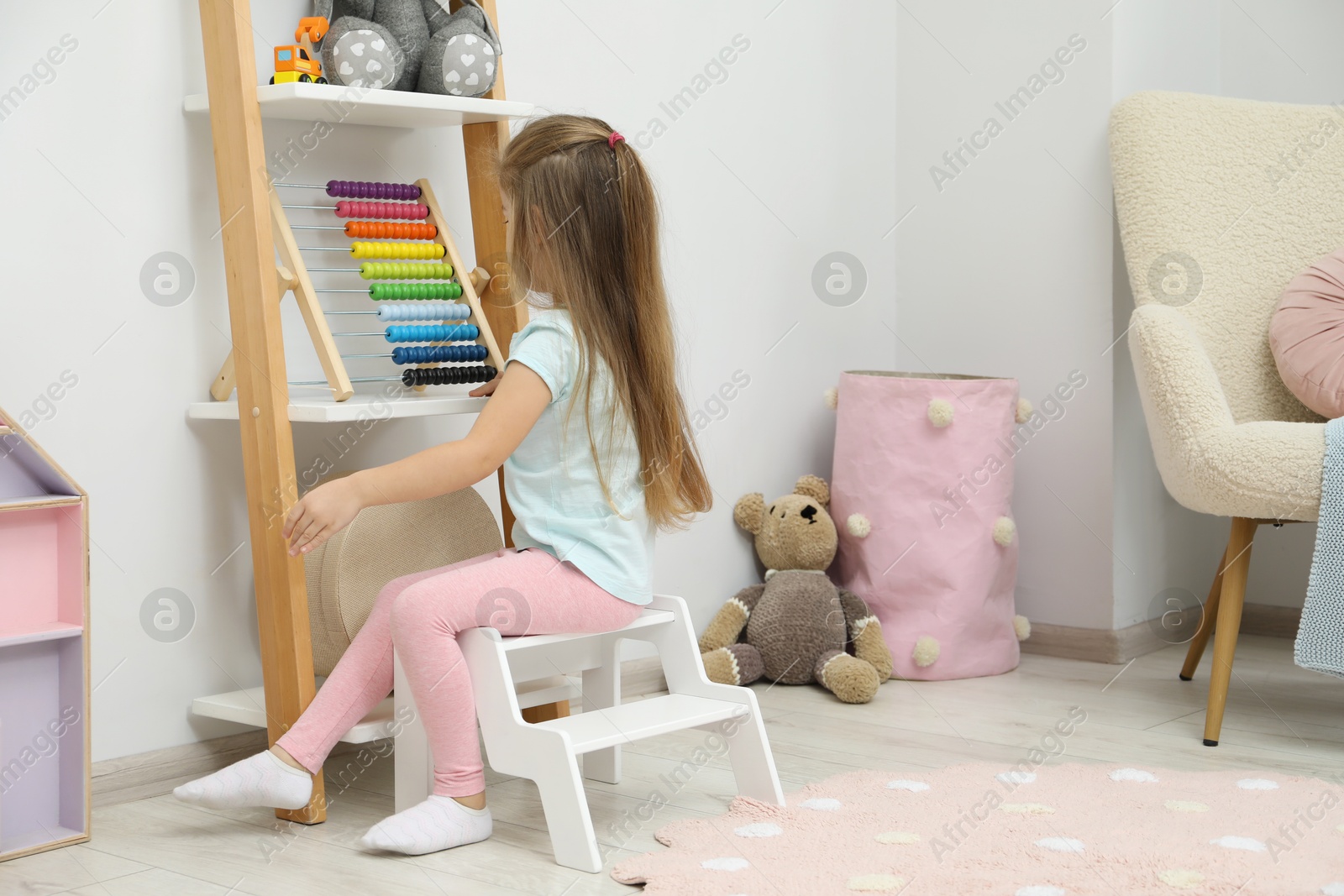 Photo of Little girl sitting on step stool near shelf with toys at home, back view