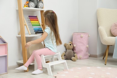 Little girl sitting on step stool near shelf with toys at home, back view