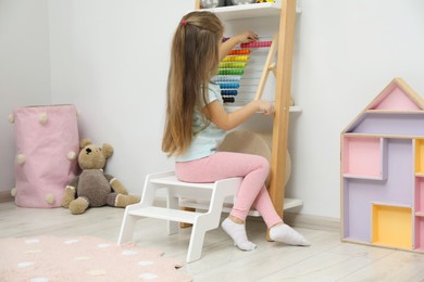 Little girl sitting on step stool near shelf with toys at home, back view