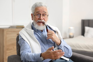 Handsome bearded man with tobacco pipe in armchair indoors