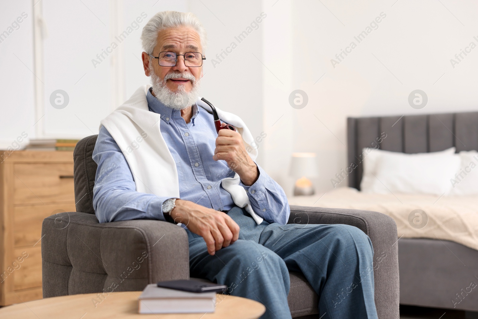 Photo of Handsome bearded man with tobacco pipe in armchair indoors