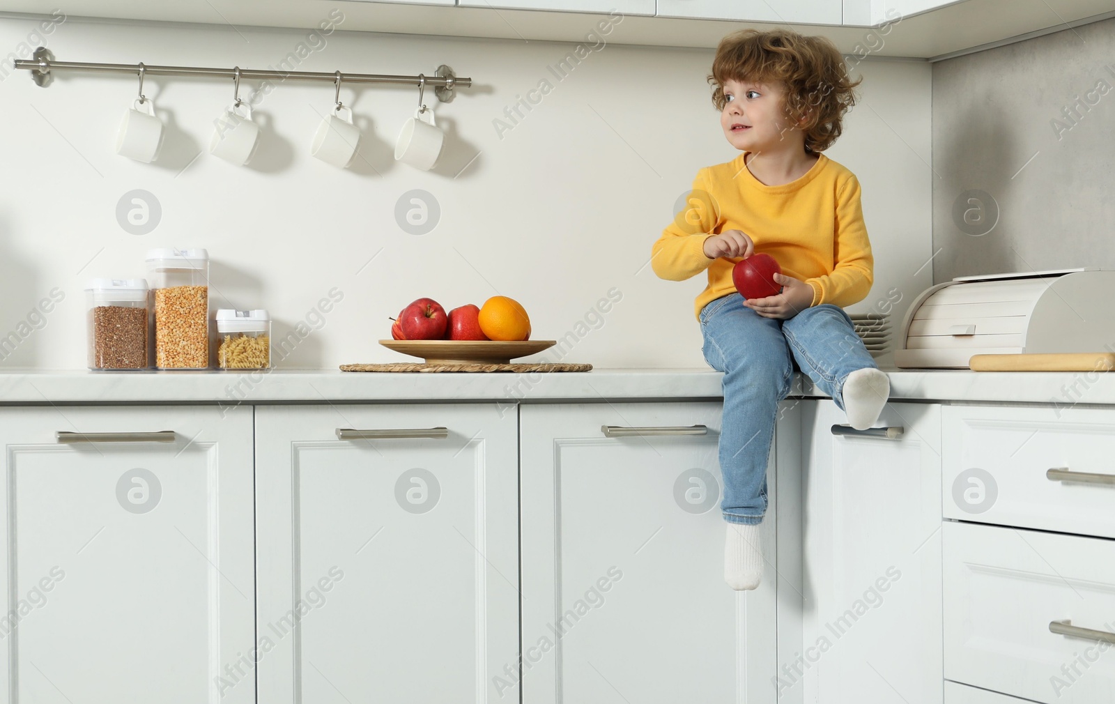 Photo of Little boy with red apple on countertop and step stool in kitchen