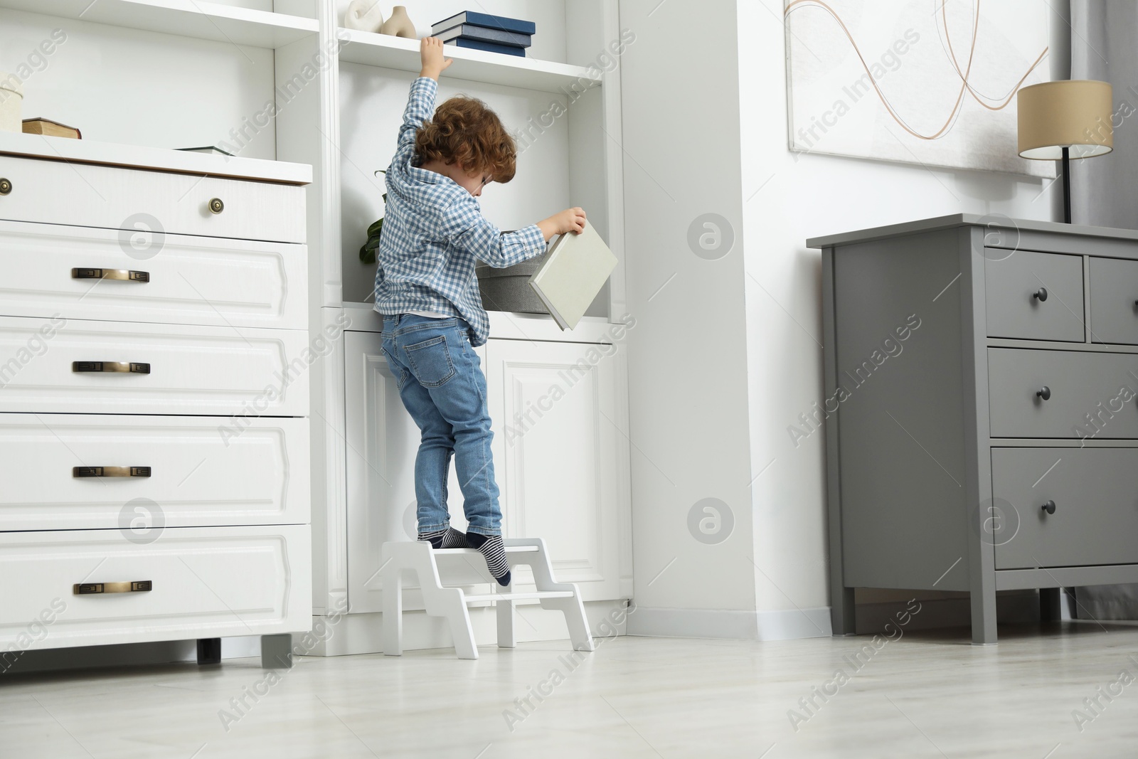 Photo of Little boy with book standing on step stool near shelving unit at home