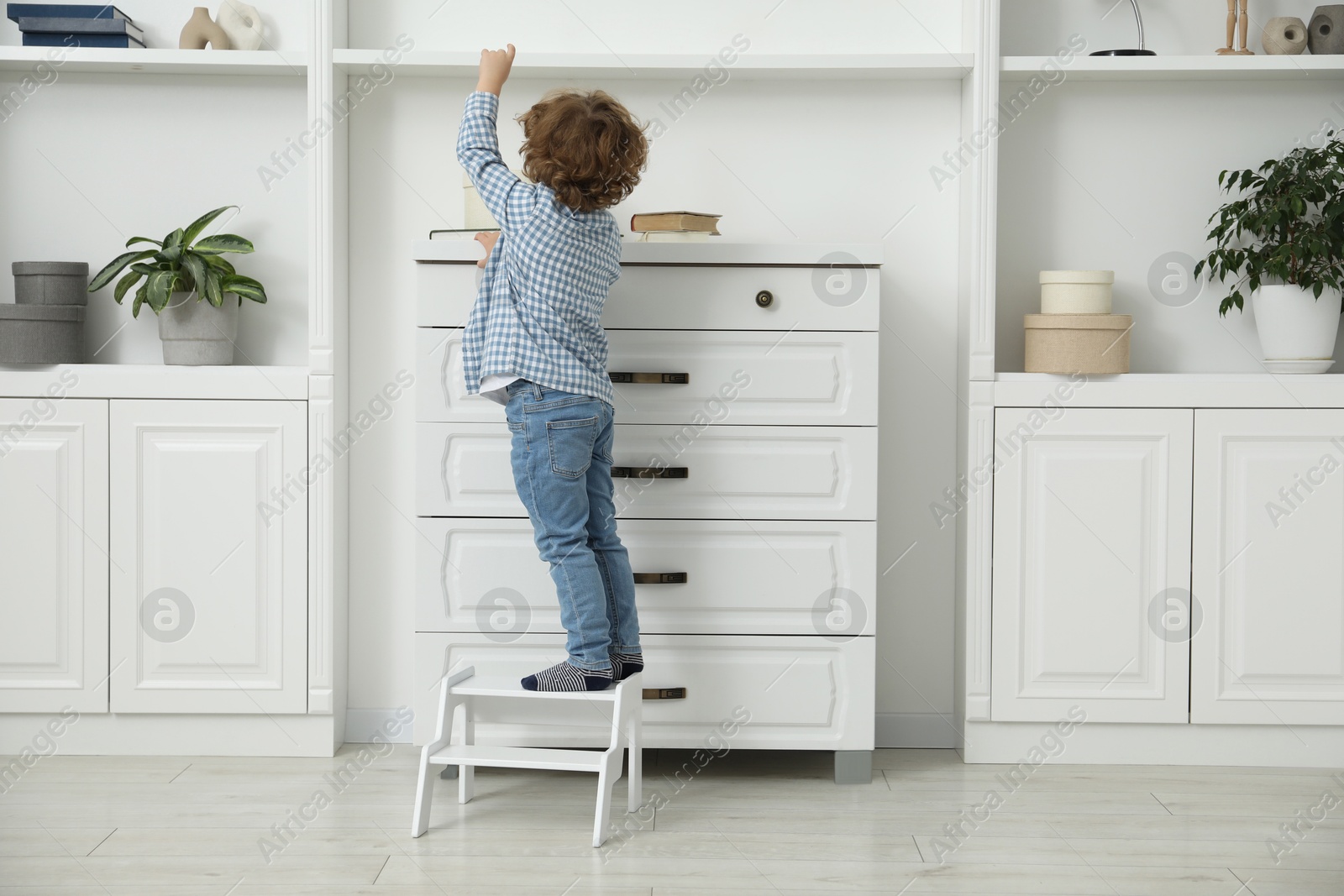 Photo of Little boy standing on step stool near chest of drawers at home