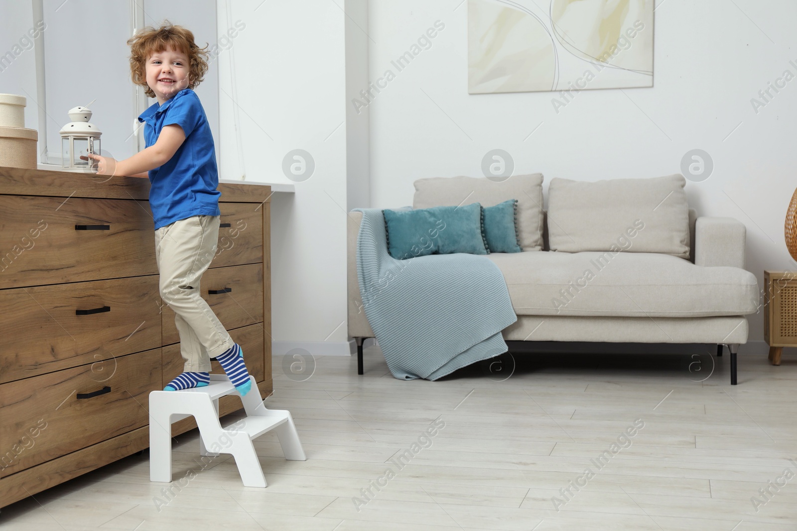 Photo of Little boy with lantern standing on step stool near chest of drawers at home