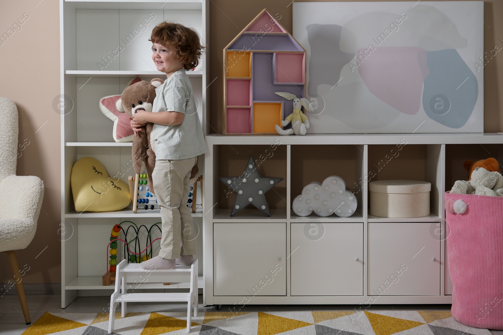 Photo of Little boy with teddy bear standing on step stool at home