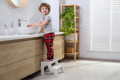 Photo of Little boy standing on step stool near bathroom vanity indoors