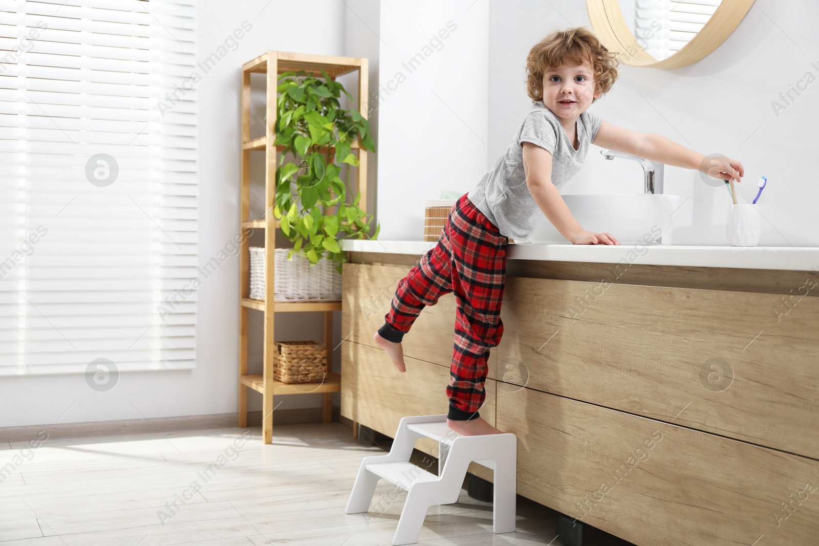 Photo of Little boy standing on step stool and reaching for toothbrush near bathroom vanity indoors