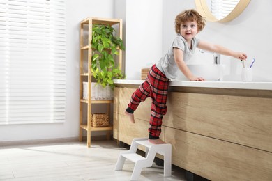 Photo of Little boy standing on step stool and reaching for toothbrush near bathroom vanity indoors