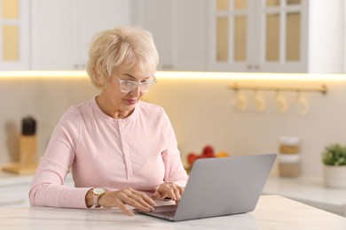 Beautiful grandmother using laptop at table in kitchen. Space for text