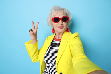 Photo of Cool grandmother showing V-sign while taking selfie on light blue background