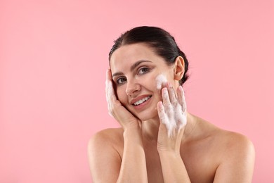 Photo of Smiling woman washing her face with cleansing foam on pink background, space for text