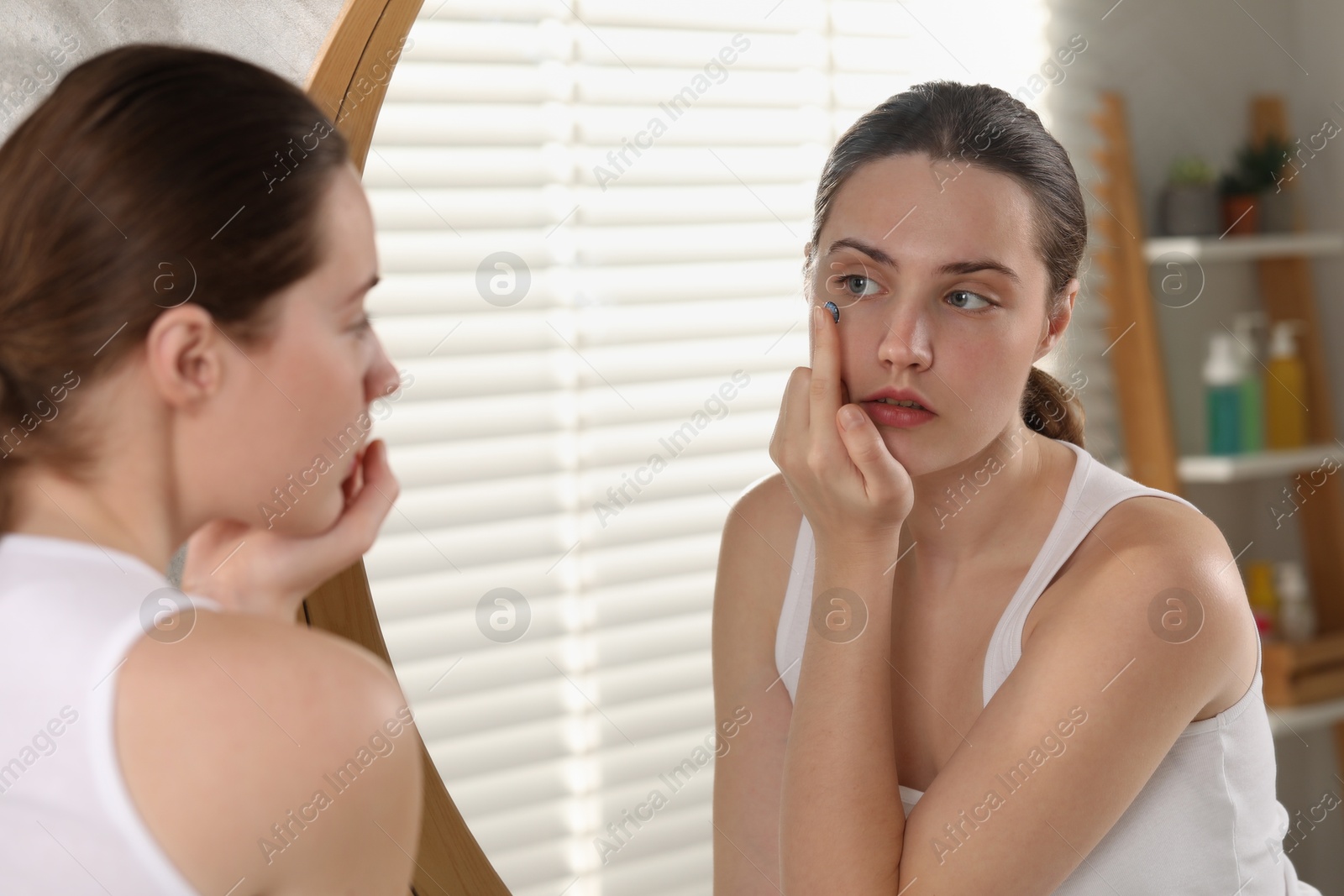 Photo of Young woman putting in blue color contact lens near mirror indoors