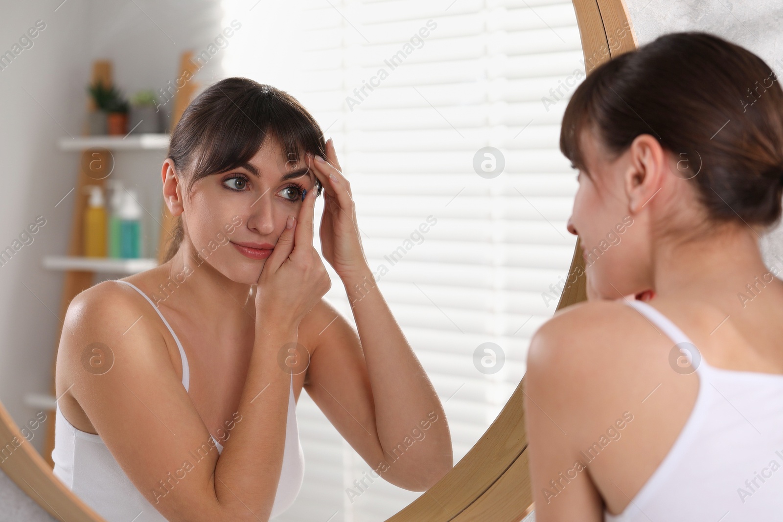 Photo of Young woman putting in color contact lens near mirror indoors