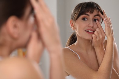 Photo of Young woman putting in red color contact lens near mirror indoors