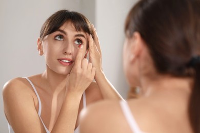 Photo of Young woman putting in red color contact lens near mirror indoors