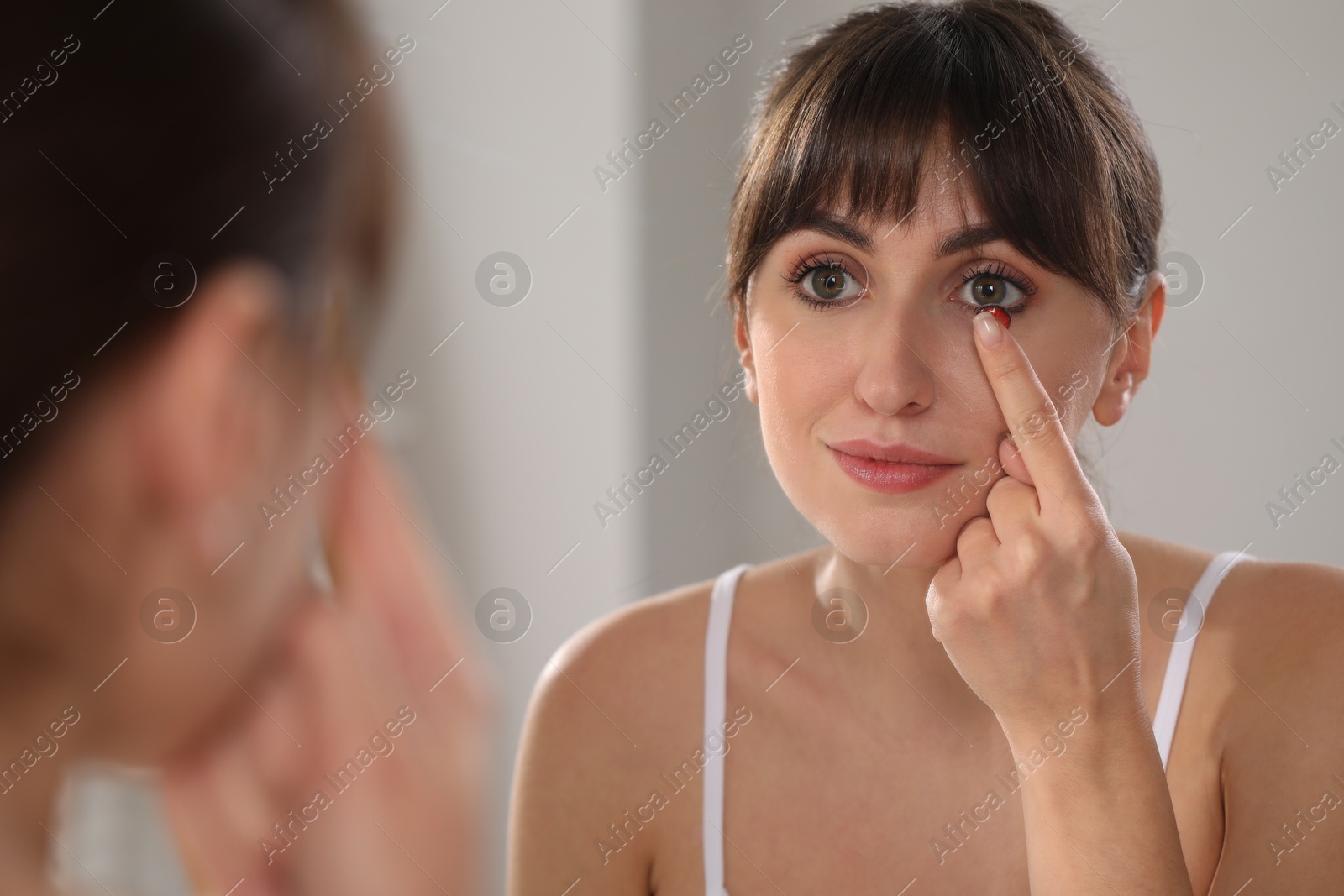 Photo of Young woman putting in red color contact lens near mirror indoors