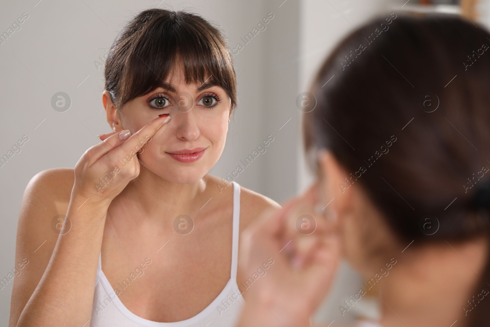 Photo of Young woman putting in red color contact lens near mirror indoors