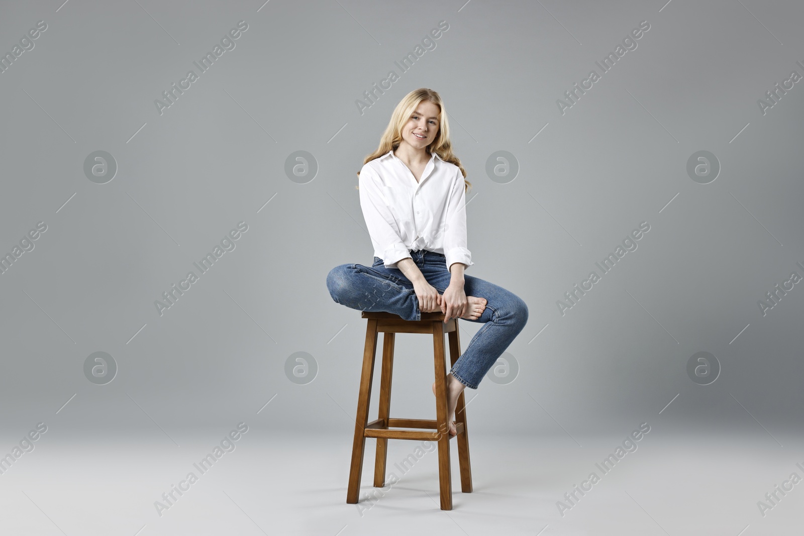 Photo of Smiling woman in stylish jeans sitting on stool against grey background