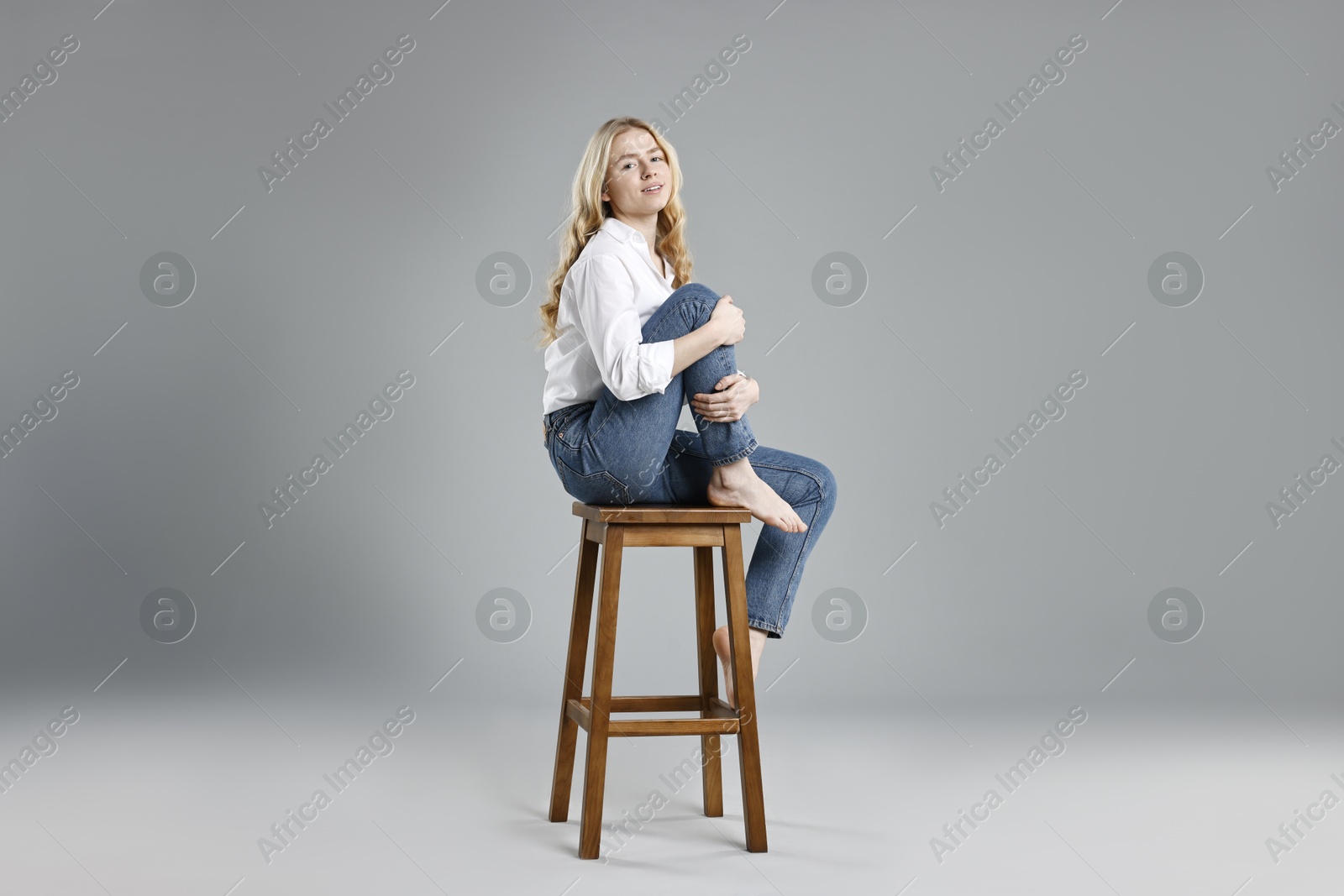 Photo of Smiling woman in stylish jeans sitting on stool against grey background