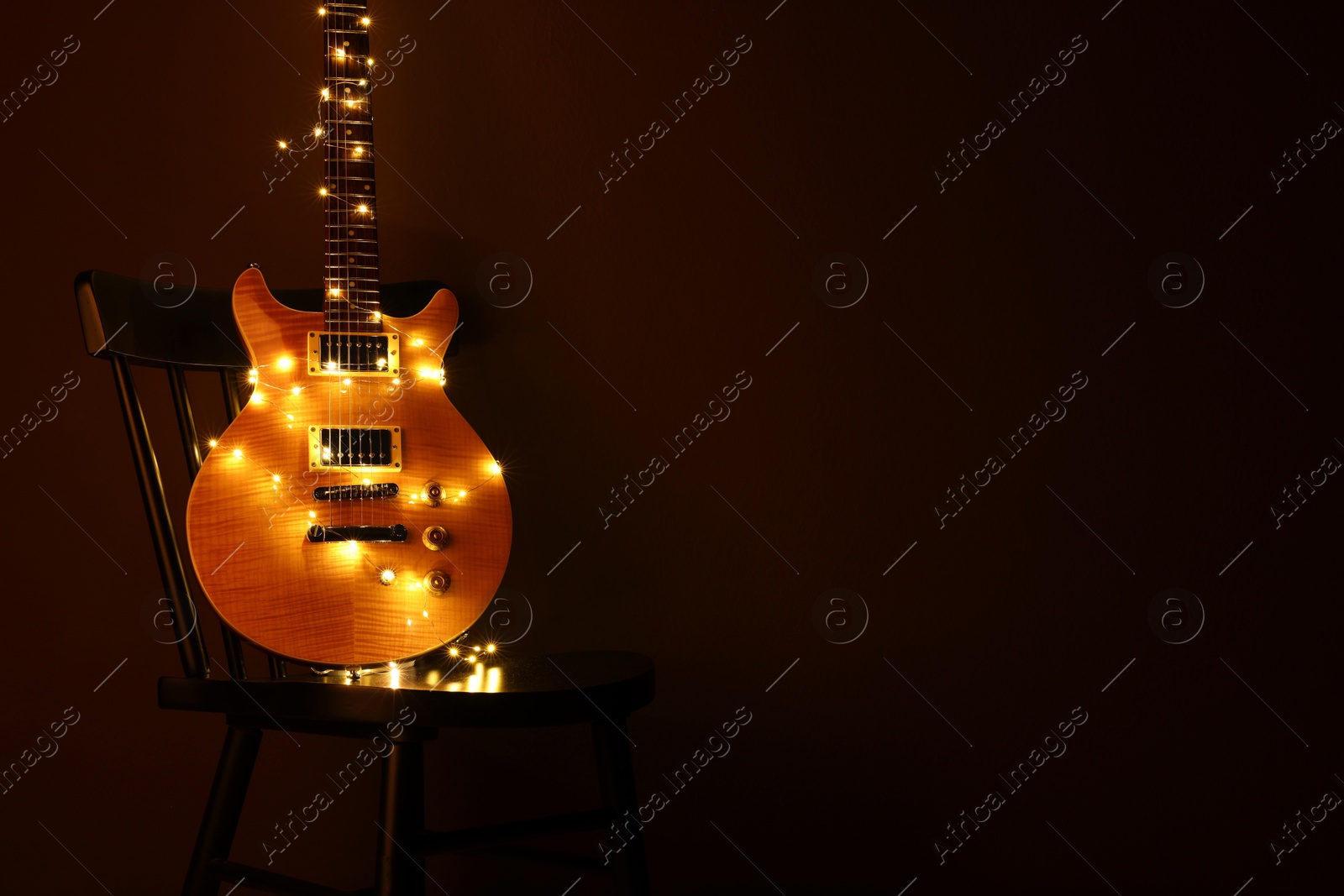 Photo of Electric guitar with Christmas lights on chair against dark background. Space for text