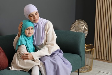 Muslim woman and her daughter sitting on sofa at home, space for text