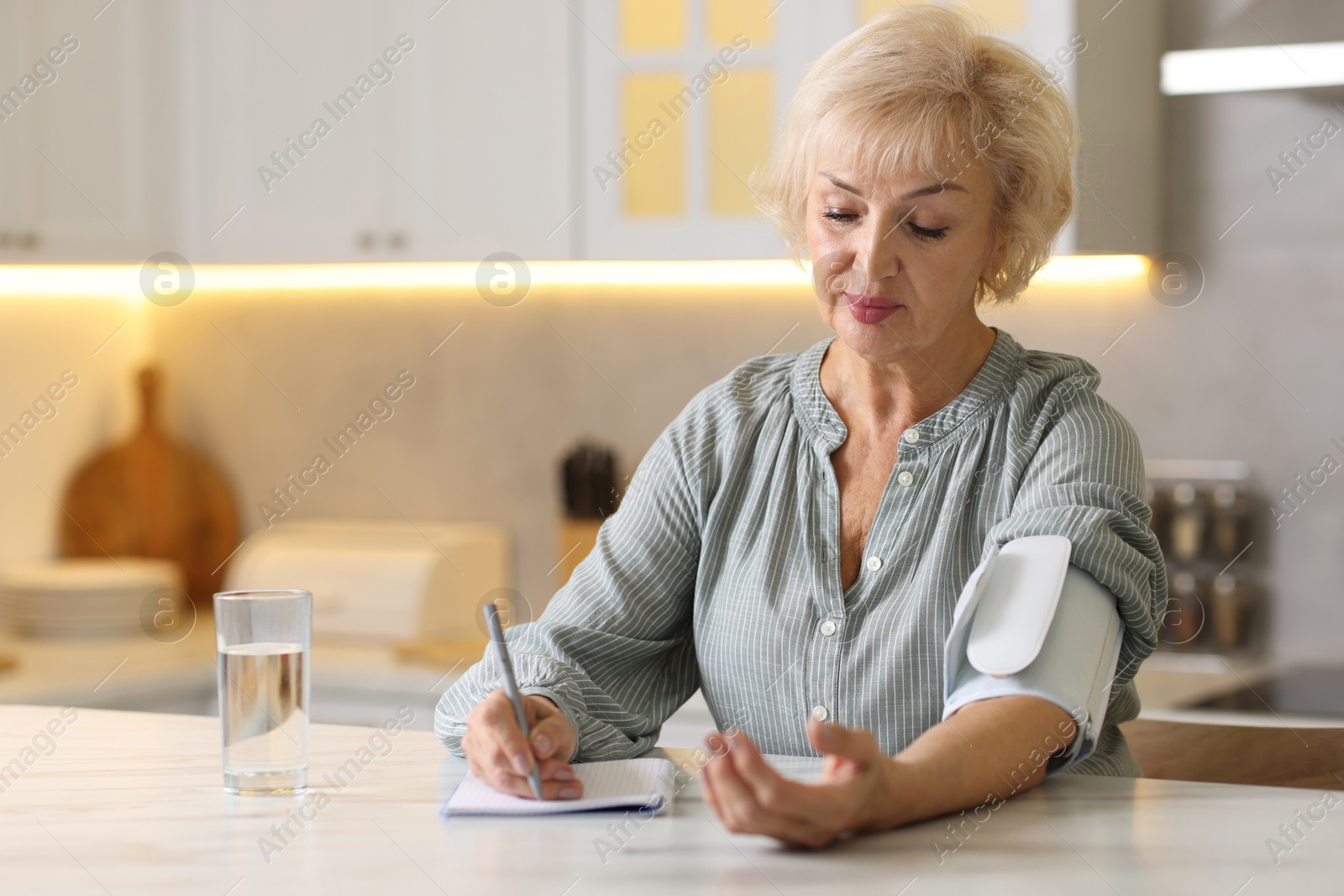 Photo of Senior woman writing results of blood pressure measurement in kitchen at home. Space for text