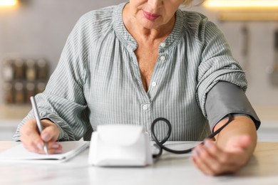 Photo of Senior woman writing results of blood pressure measurement in kitchen at home, closeup