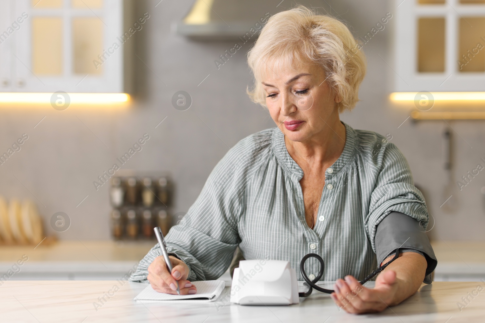 Photo of Senior woman writing results of blood pressure measurement in kitchen at home. Space for text