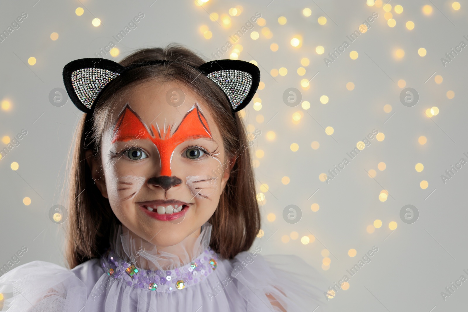 Photo of Cute girl with painted face and ears as cat on grey background with blurred lights, space for text