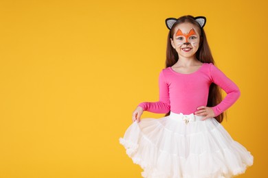 Photo of Cute girl with painted face and ears as cat on orange background, space for text