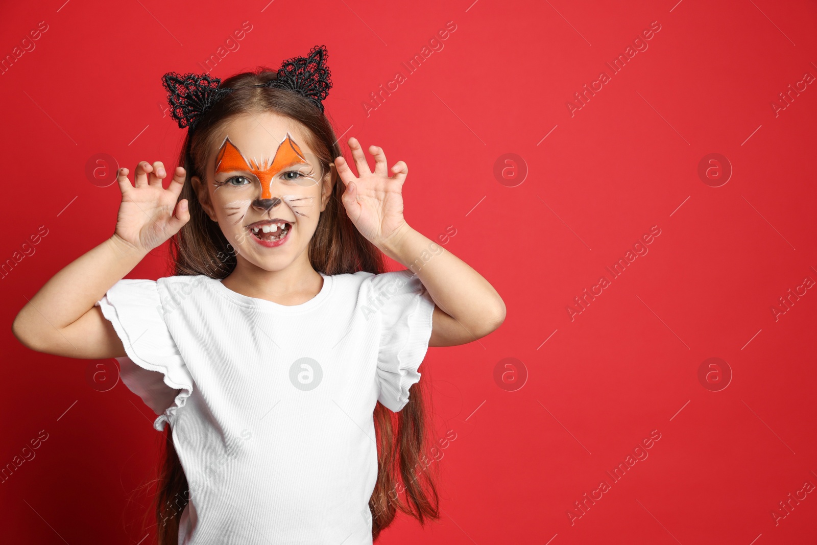 Photo of Cute girl with painted face and ears as cat on red background, space for text