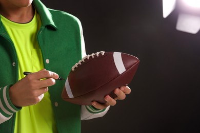 Photo of Professional player signing autograph on American football ball against dark background, closeup. Space for text